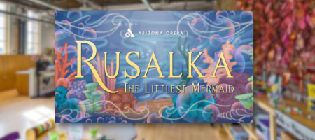 A photo of the room in Children's Museum of Phoenix with several desks with arts and craft supplies. The graphic for the Arlyn M. Brewster OperaTunity program's production of Rusalka: The Littlest Mermaid is in the center of the photo with a box shadow around it.